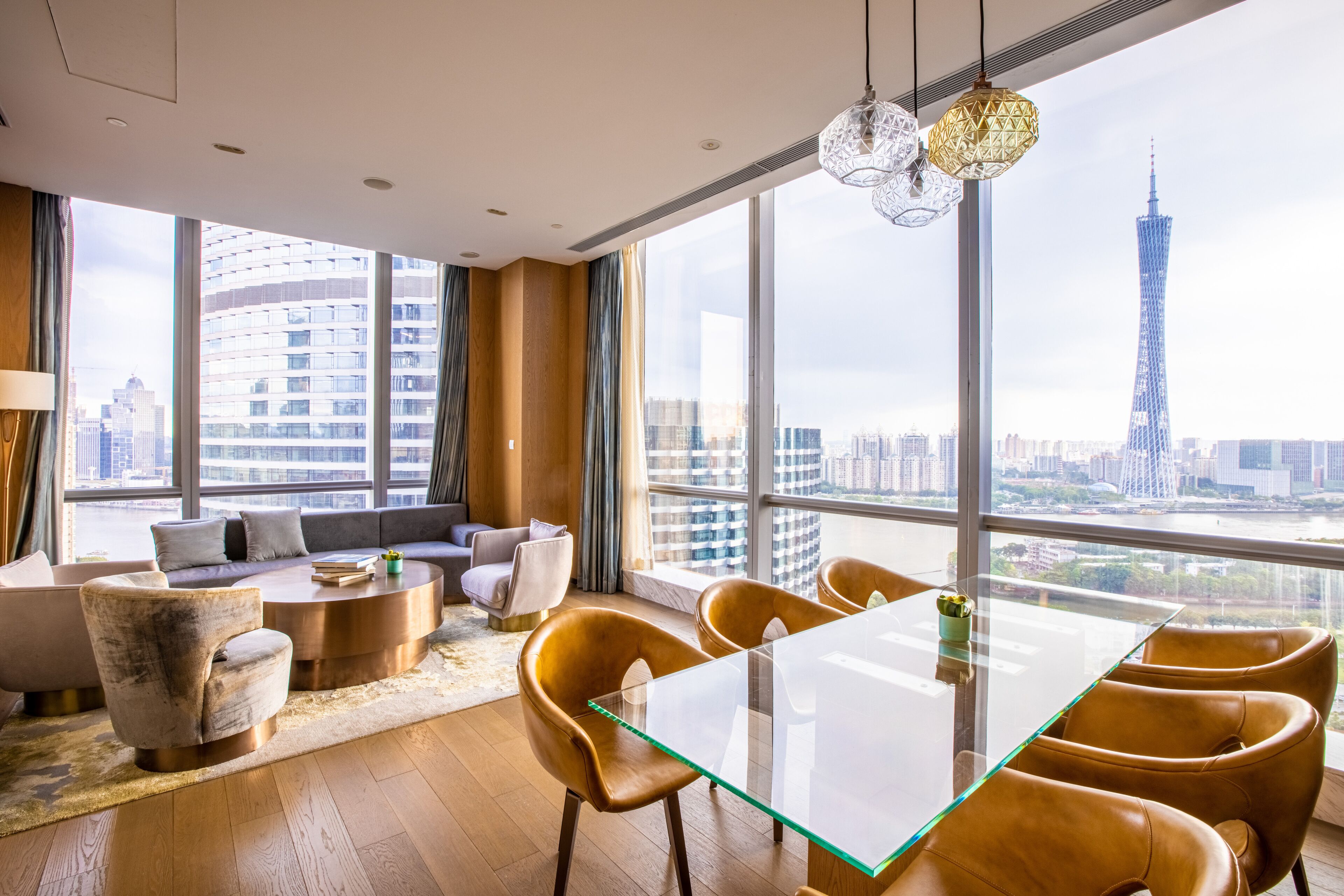 Una habitación de hotel moderna con un ventanal grande, una mesa de cristal, sillas de cuero, un sofá y vistas a un paisaje urbano que incluyen una torre alta.