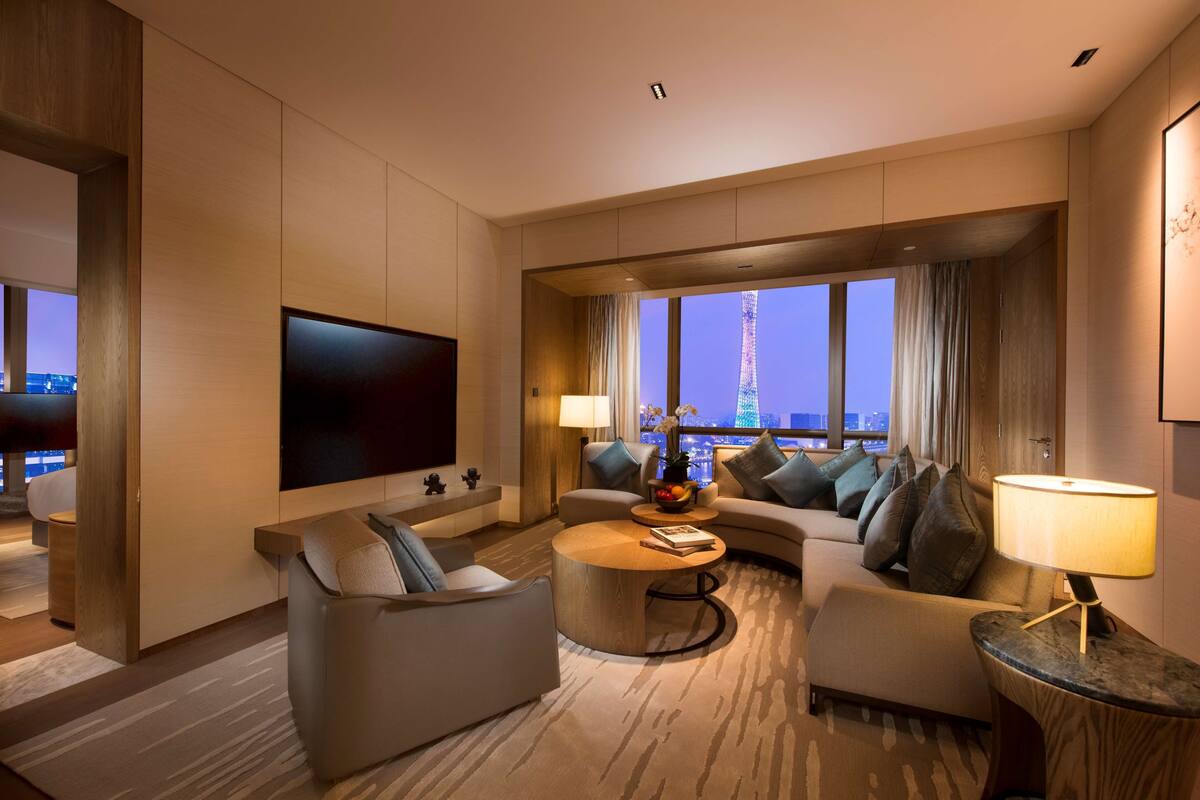 A modern hotel room with a large flat-screen TV, a sofa, a coffee table, and a view of the cityscape.
