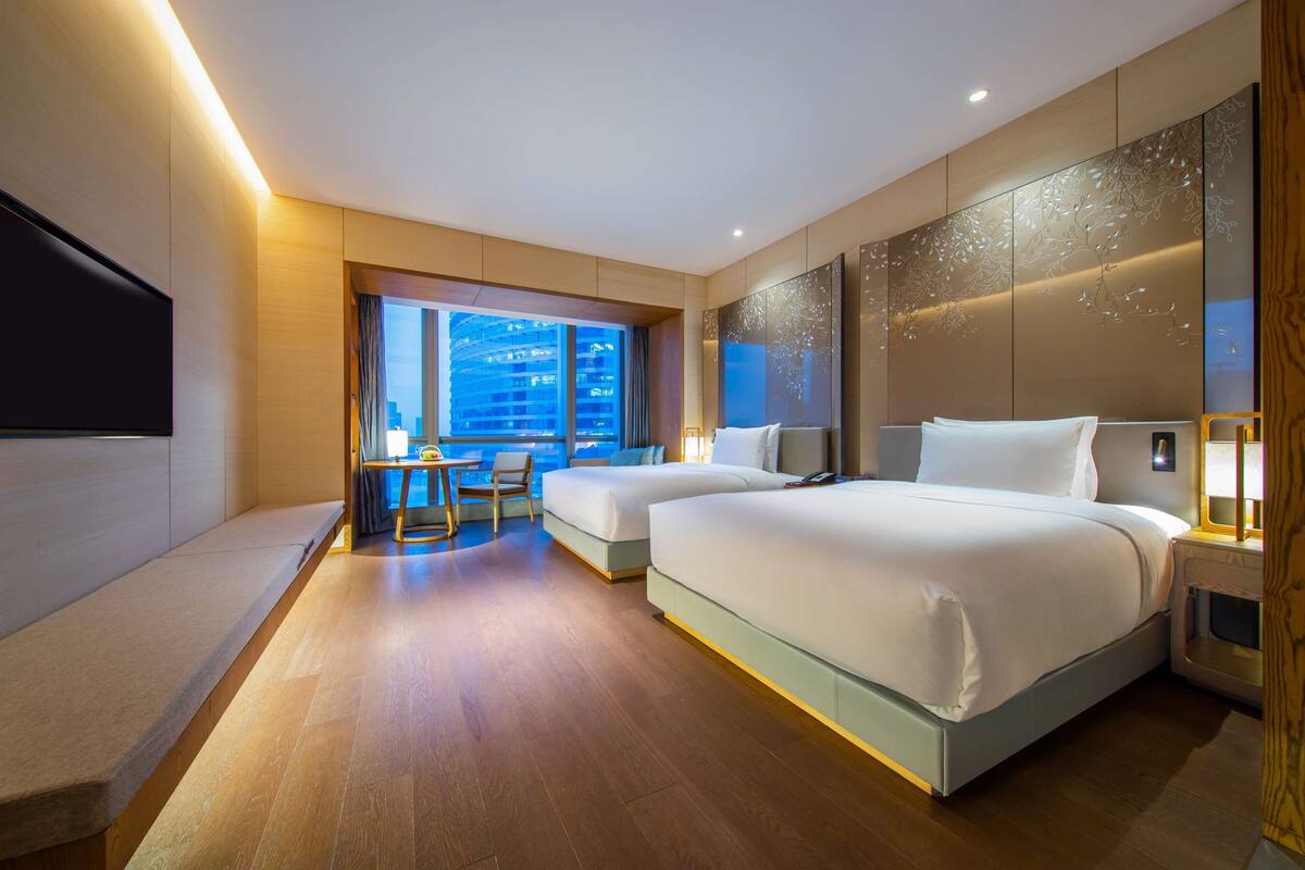 A hotel room with two beds, a desk, and a large window offering a city view.