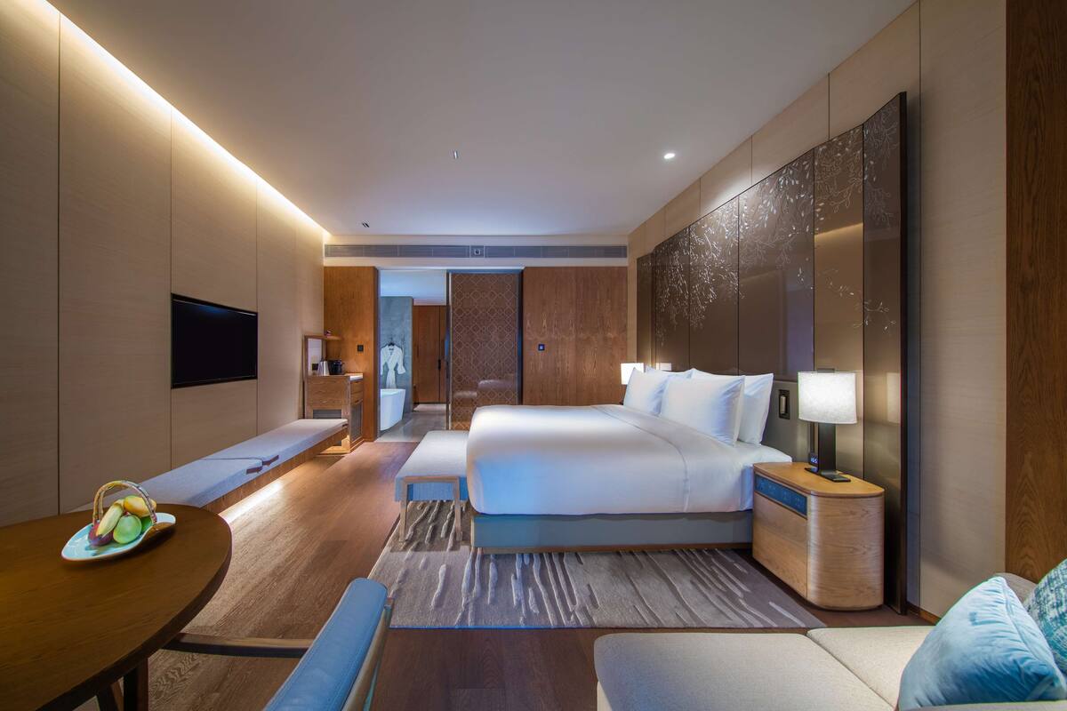 A modern hotel room with a large bed, bedside tables, a flat-screen TV, a dining table with fruit, and a view of the hallway.