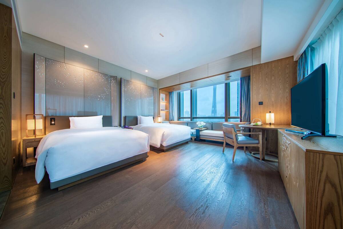 A modern hotel room with two beds, a desk, a TV, and large windows.