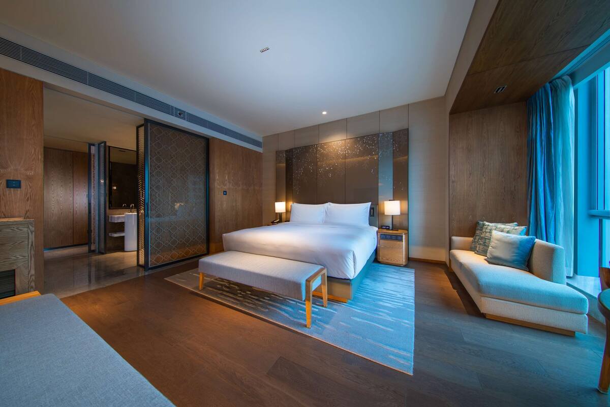 A modern hotel room with a large bed, bedside tables, a sofa, and a view of the city.