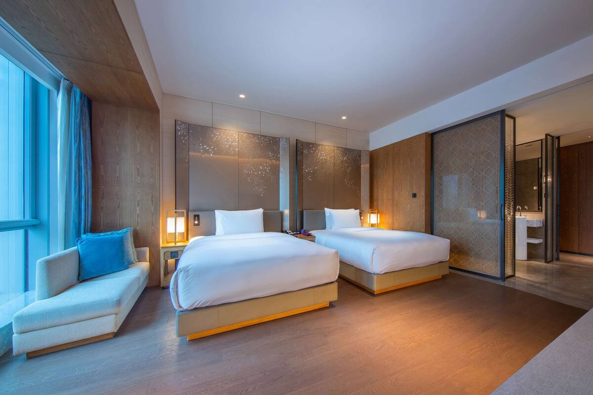 A modern hotel room with two beds, a sofa, and a large window.