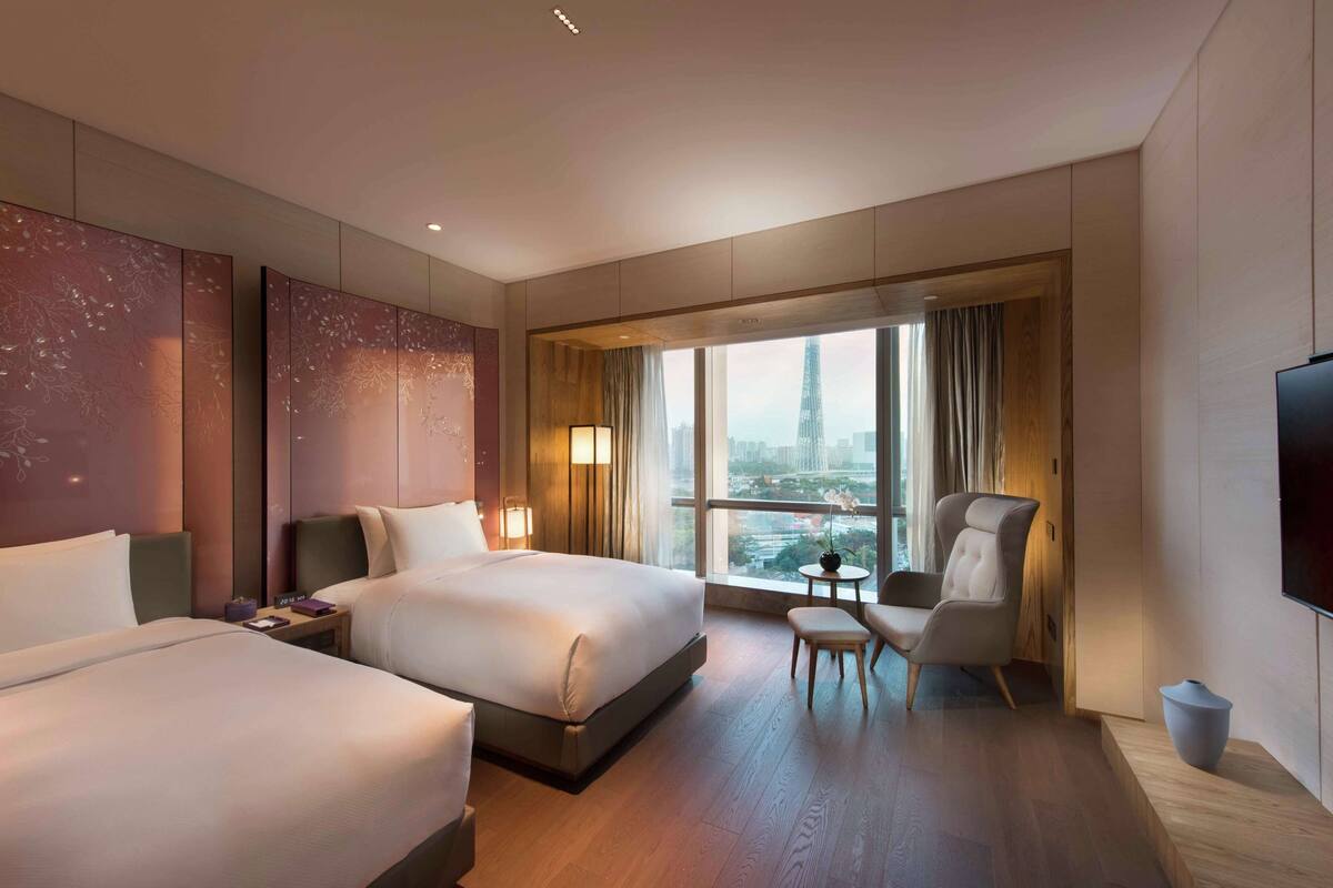 A hotel room with two beds, a seating area, a TV, and a large window with a city view.