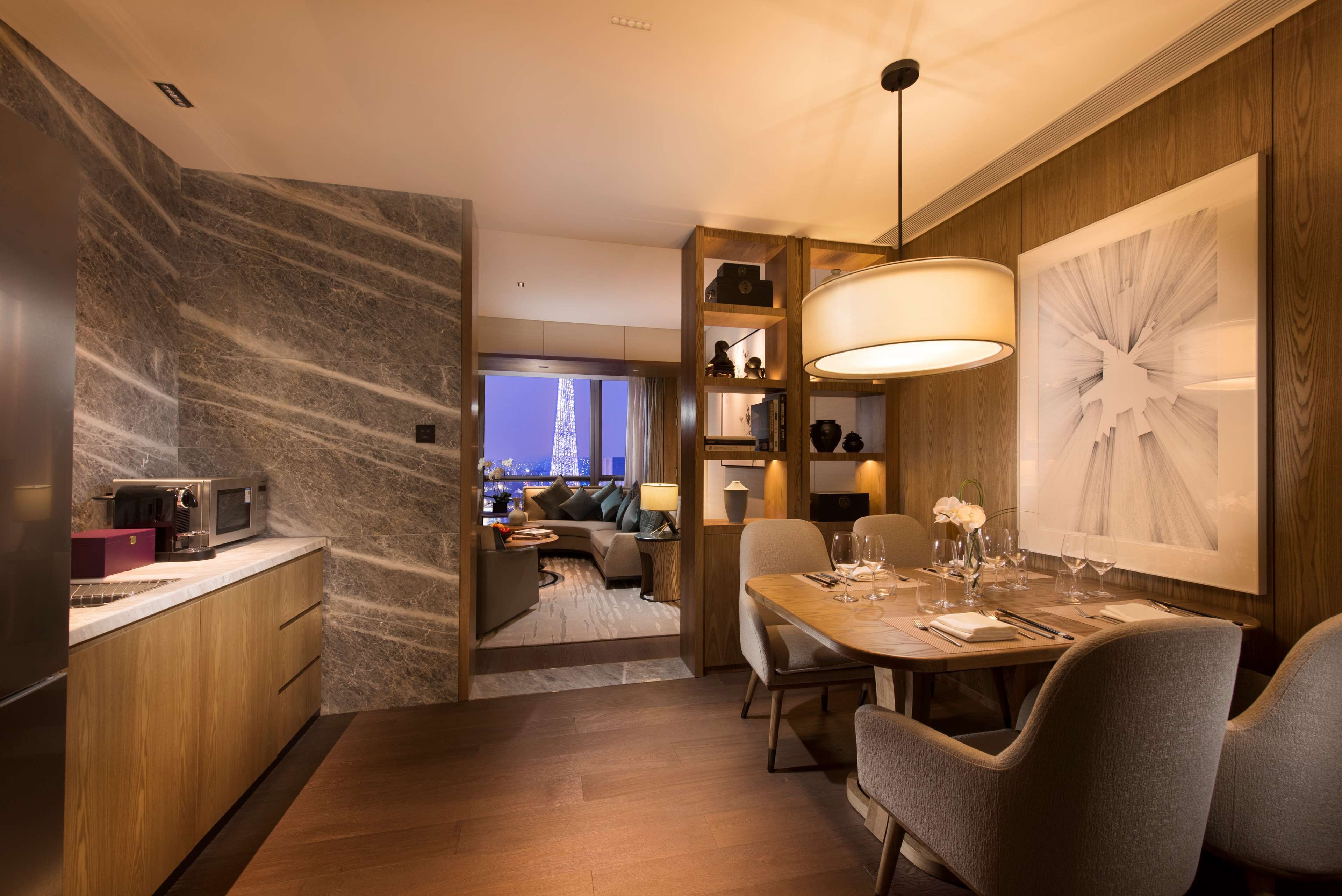 A modern hotel room with a dining area, kitchen, and a view of the cityscape.