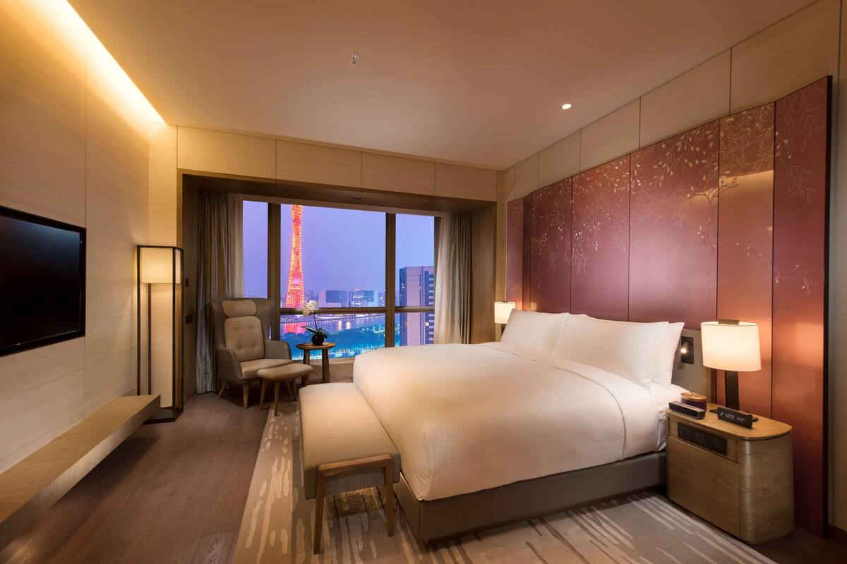 A hotel room with a large bed, a TV, a desk with a lamp, and a view of a cityscape.
