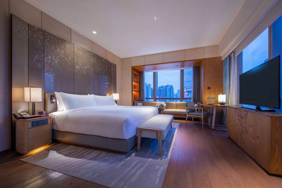 A modern hotel room with a large bed, bedside tables, a desk, a television, and a view of the cityscape.