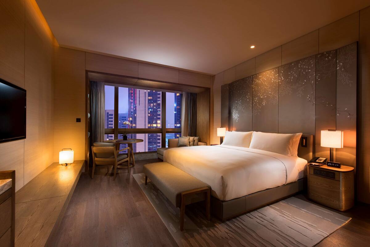 A modern hotel room with a large bed, a desk, a chair, a bench, a TV, and a view of the cityscape.