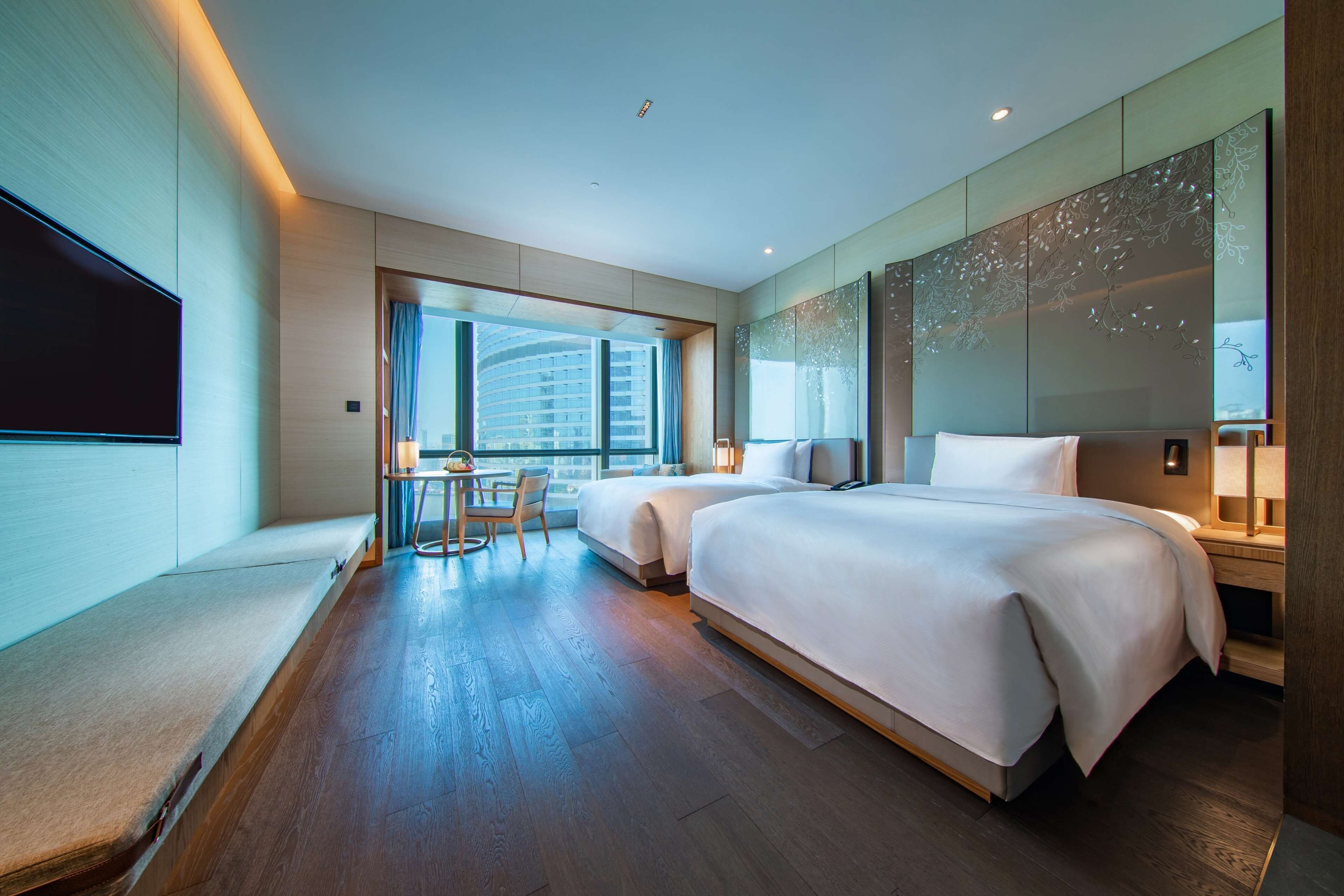 A modern hotel room with two beds, a large window with a view of a building, a flat-screen TV, and a small table with chairs.