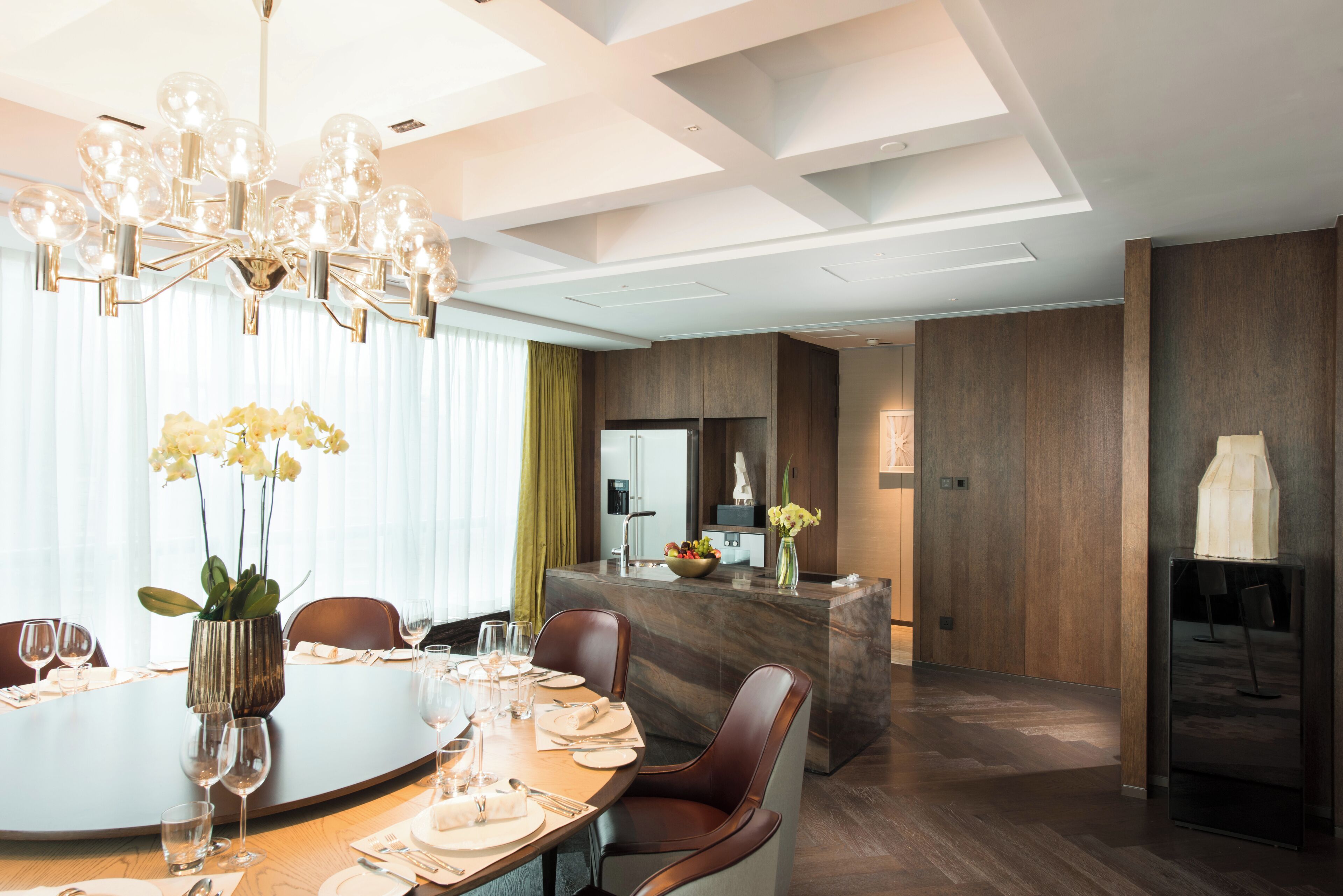 Uma área de jantar moderna com uma mesa redonda posta para seis pessoas, um candelabro e vista para a cozinha ao fundo.