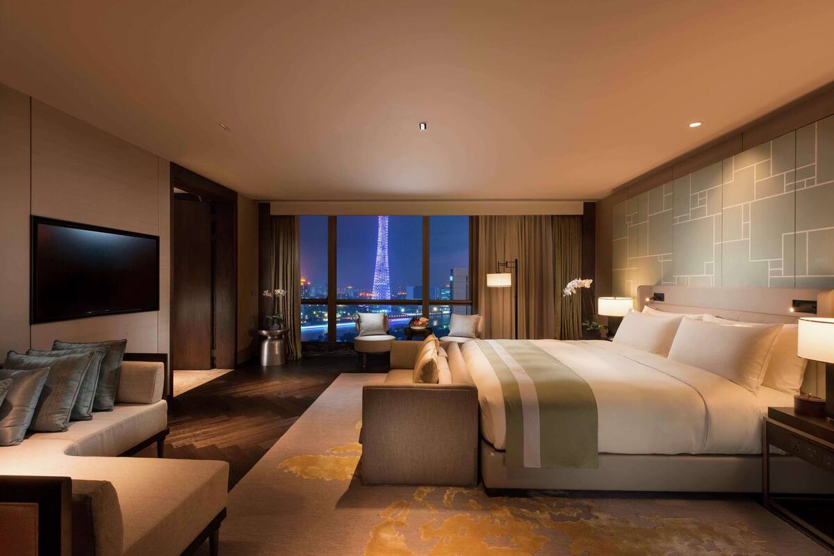 A modern hotel room with a large bed, a sofa, a TV, and a view of the cityscape at night.