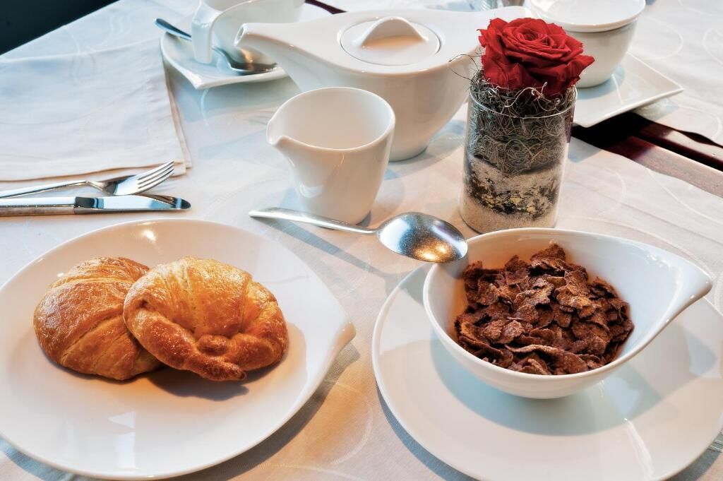 A table set with croissants, a bowl of cereal, a teapot, a cup, and a vase with a rose.