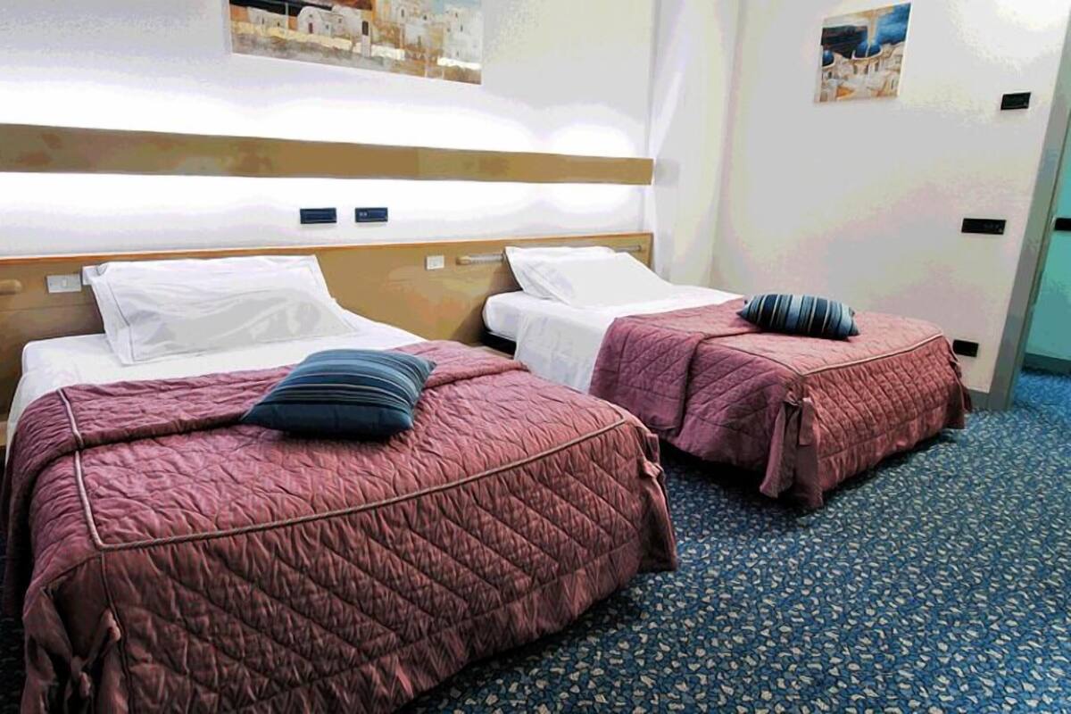 Une chambre d’hôtel avec deux lits, chacun recouvert d’une courtepointe rose matelassée et agrémenté d’oreillers. La chambre est dotée d’une moquette bleue et d’une tête de lit en bois.