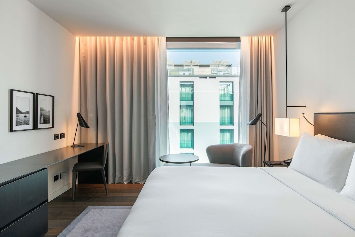 A hotel room with a bed, desk, chair, lamp, and a view of buildings through the window.