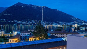 A modern building with a glass conservatory, a courtyard with outdoor seating, and a view of a lake and mountains in the background. - Hilton Lake Como (Como)