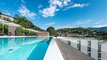 Ein moderner Infinity-Pool mit klarem, blauem Wasser, umgeben von einer Holzterrasse und Glasgeländern, mit Blick auf einen Hügel mit üppiger Vegetation und blauem Himmel.