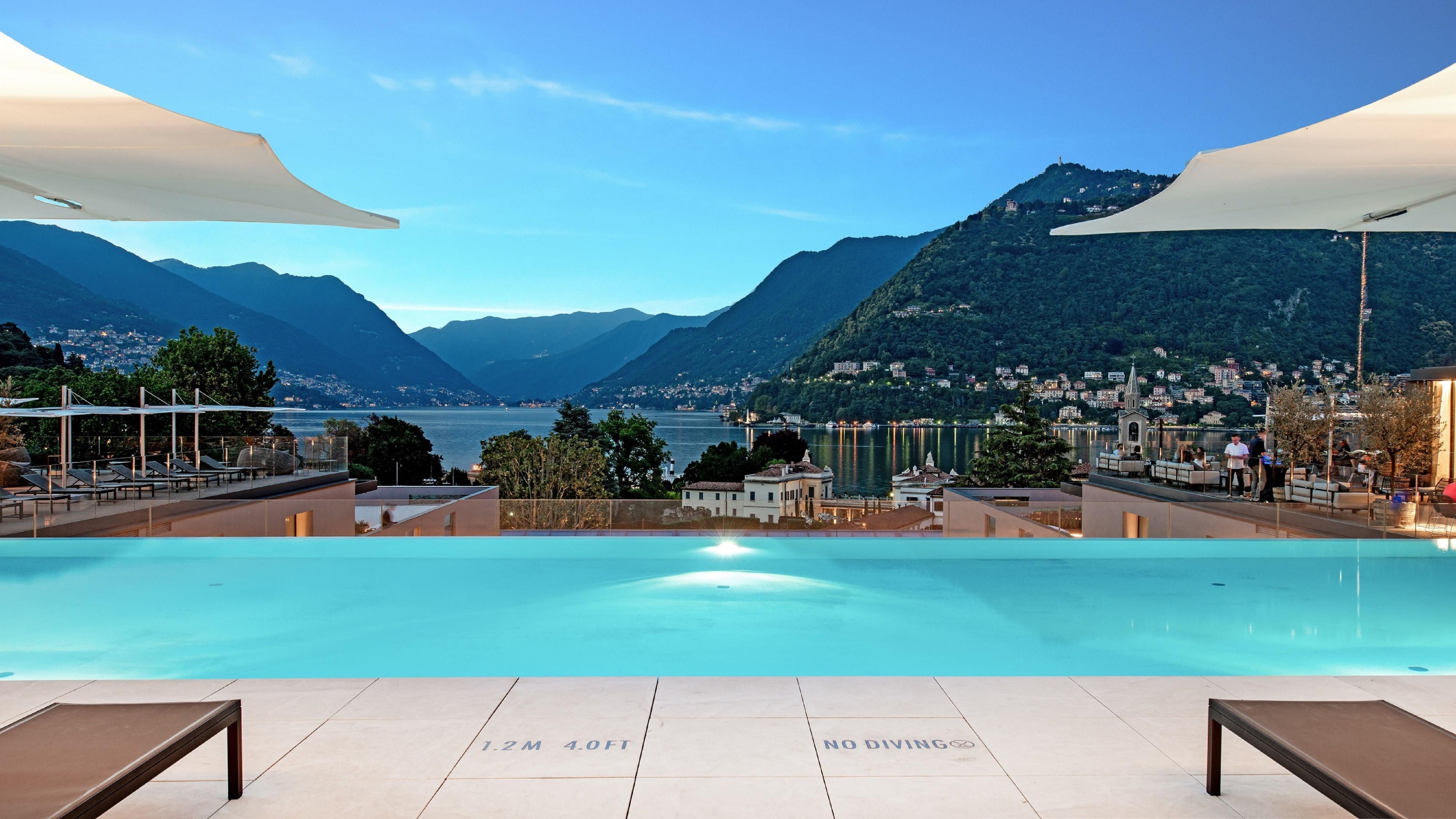 Un espace piscine avec vue sur les montagnes et un lac, des chaises longues et un panneau indiquant la profondeur de la piscine et interdisant les plongeons.