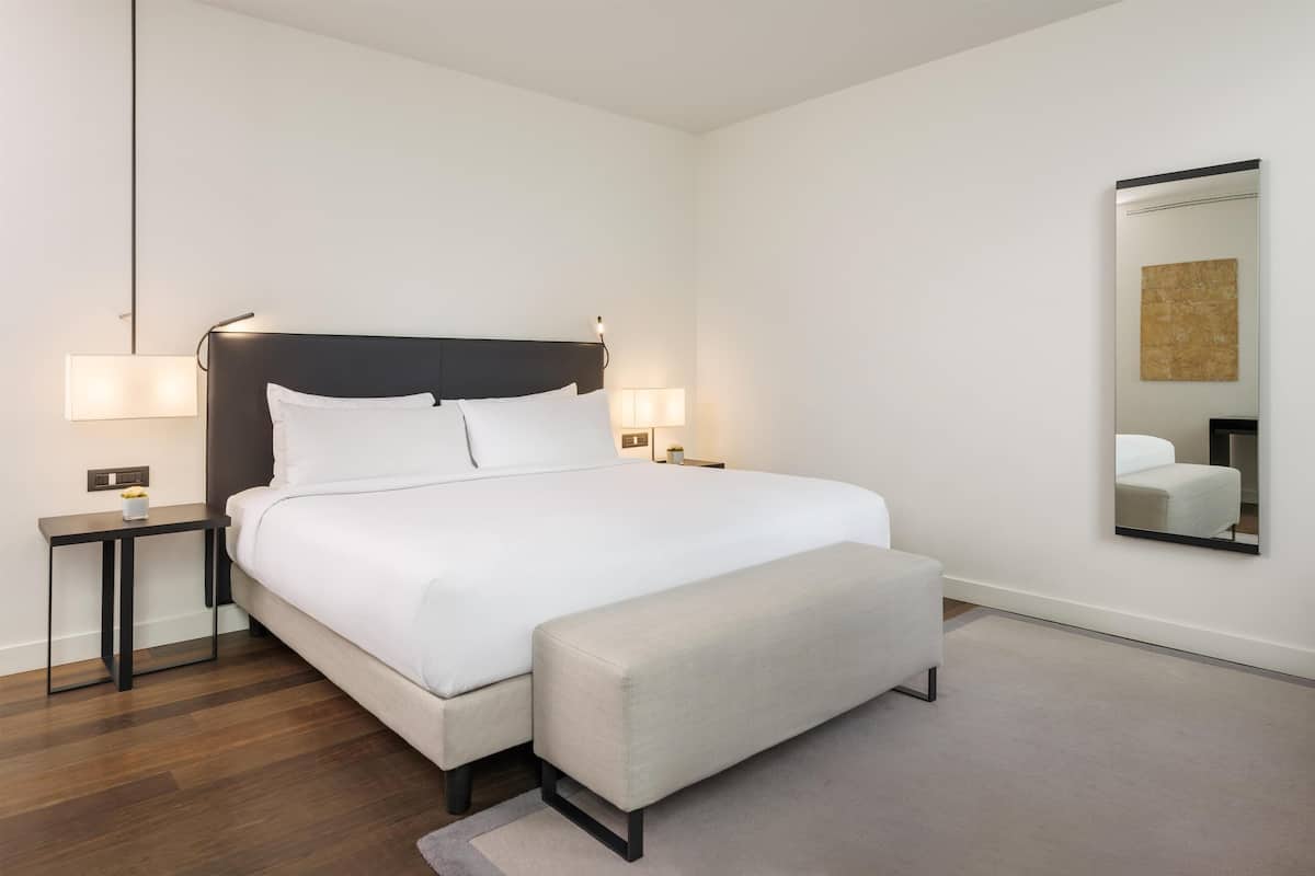 Une chambre d’hôtel moderne avec un grand lit, des lampes de chevet, un banc et un miroir au mur.