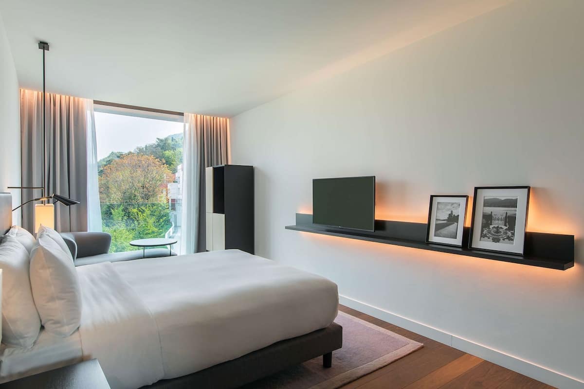 Une chambre d’hôtel moderne avec un grand lit, une télévision, une étagère avec des photos et une vue sur l’extérieur.