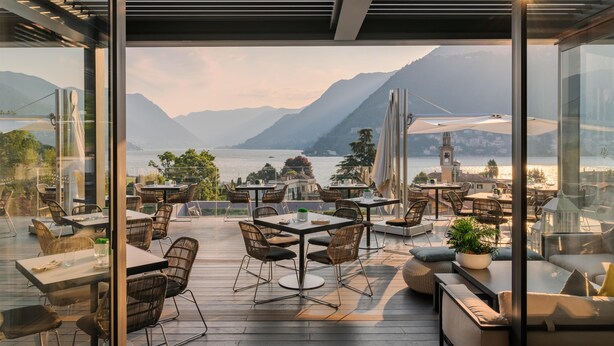 An outdoor terrace with tables and chairs, overlooking a lake and mountains. - Hilton Lake Como (Como)
