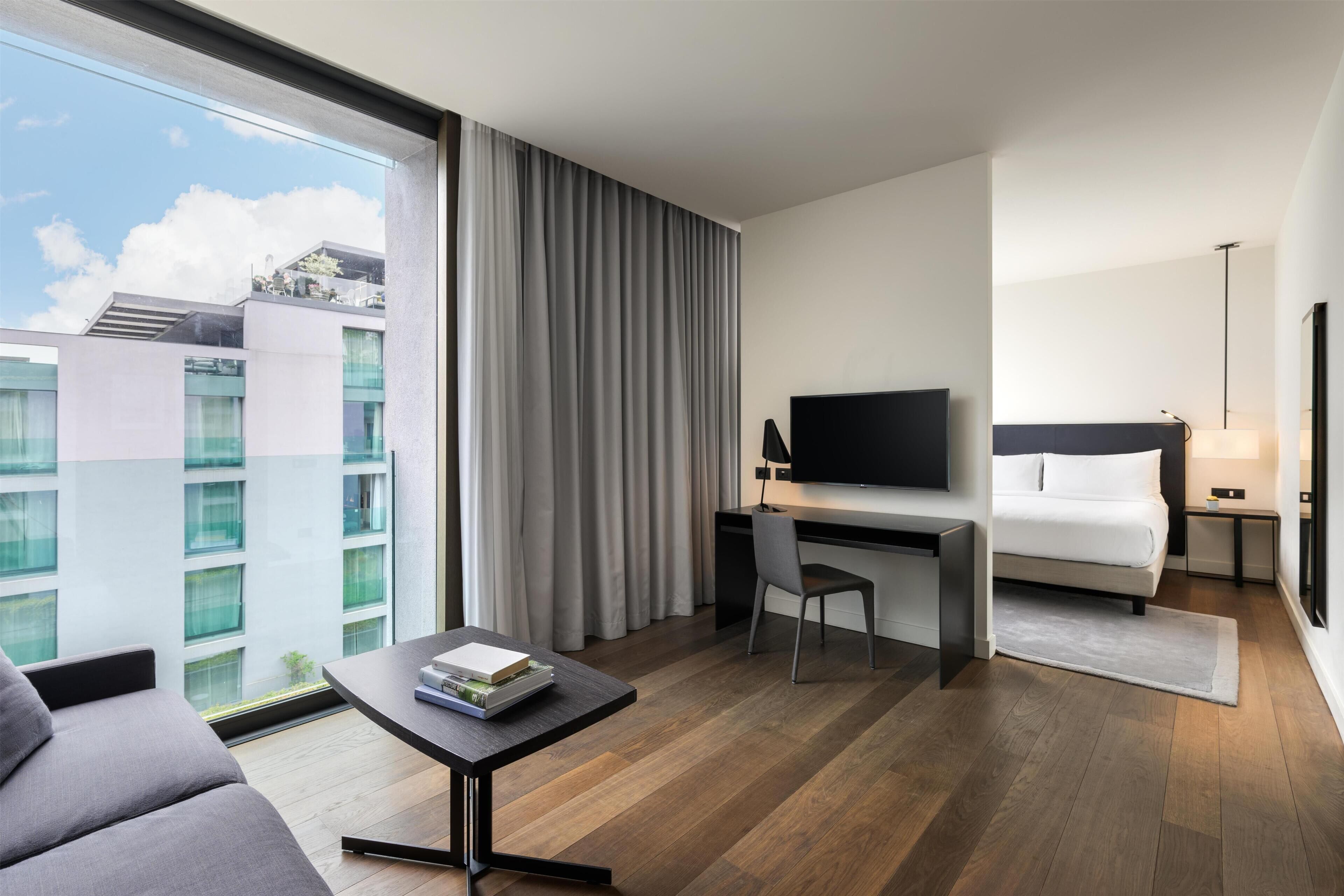 A hotel room with a bed, a desk with a TV, a sofa, and a view of buildings.
