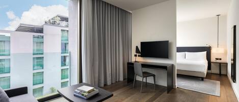 A hotel room with a bed, a desk with a TV, a sofa, and a view of buildings.