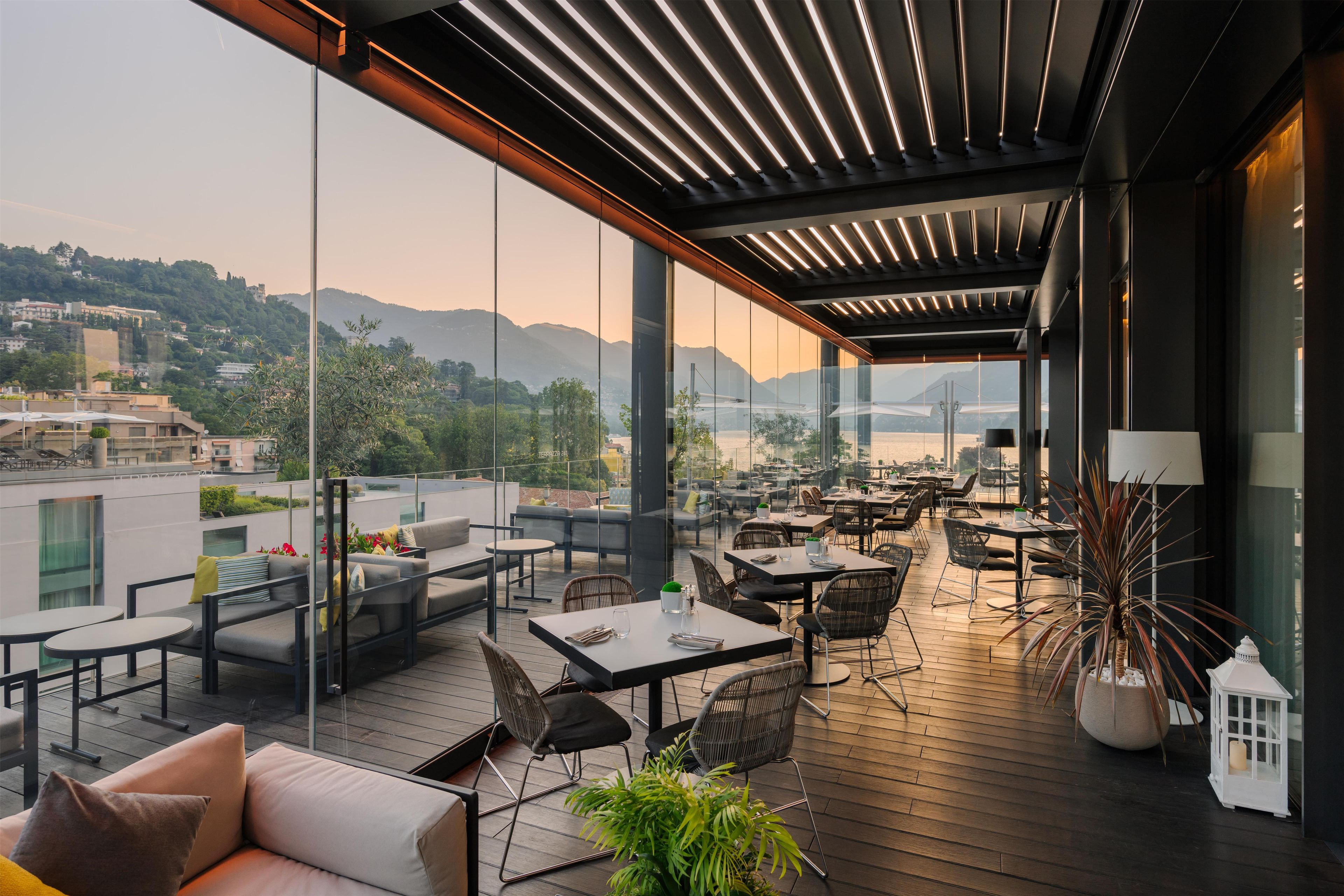 A modern outdoor dining area with glass walls, wooden flooring, and a view of a cityscape and mountains.