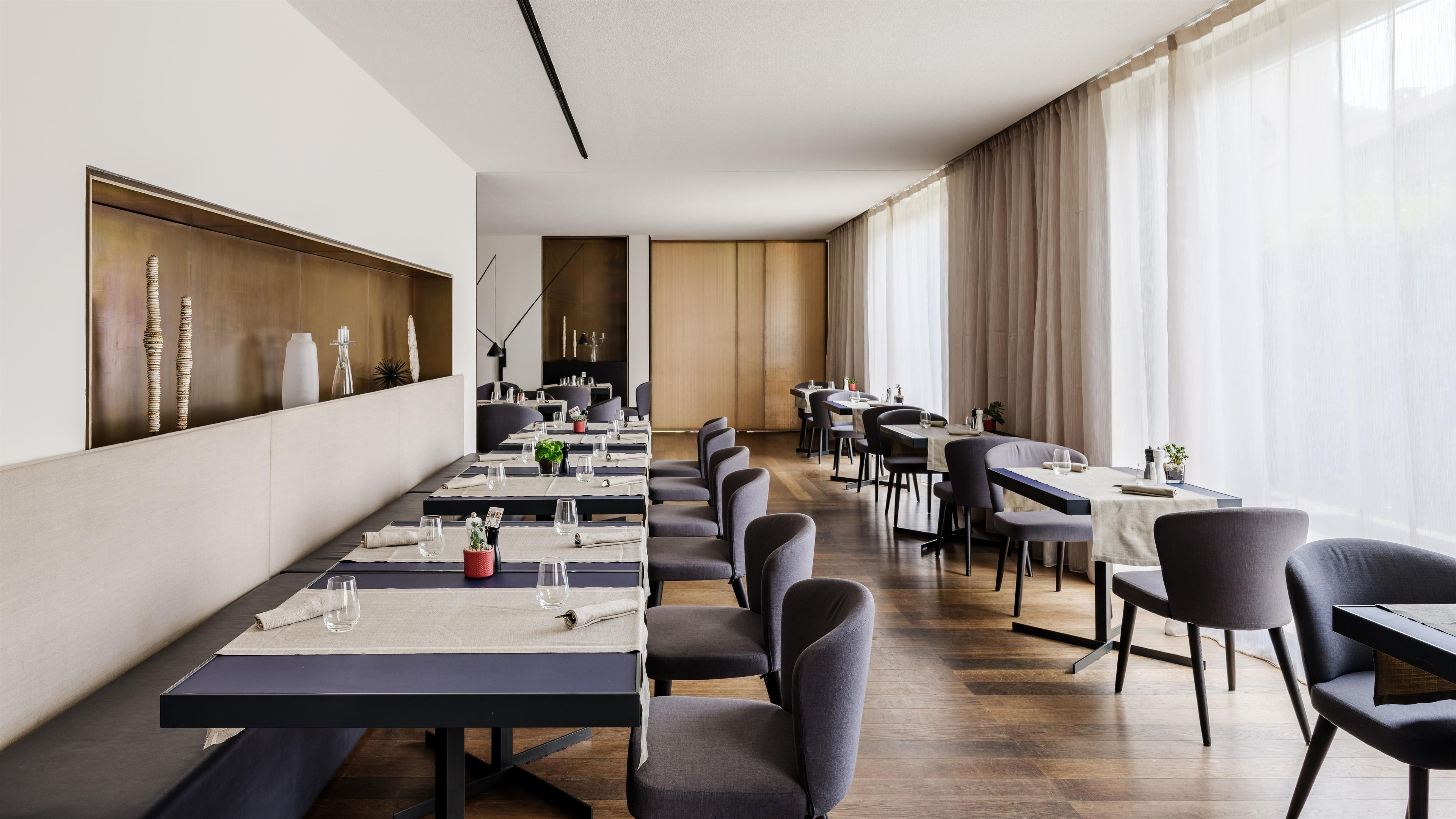 Un área de comedor moderna con mesas largas, sillas y un amplio ventanal con cortinas.