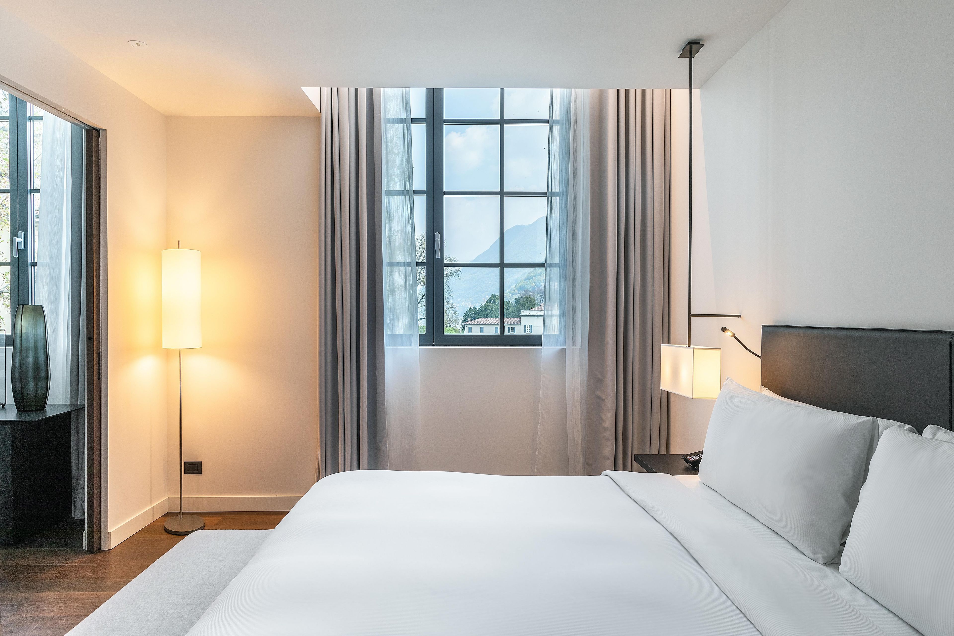 A hotel room with a large bed, a bedside table, a lamp, a view of the outdoors through a window, and a door leading to another room.