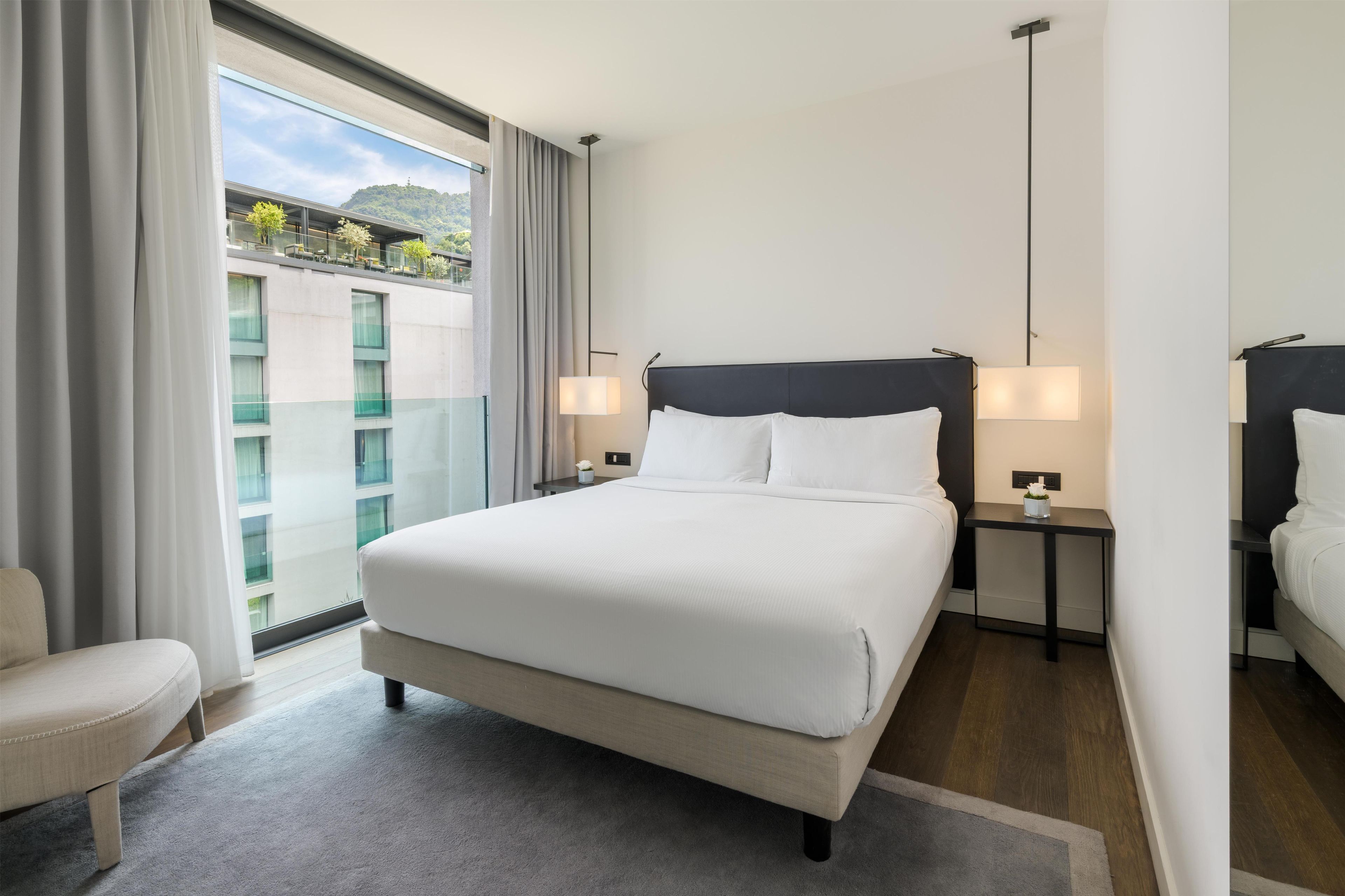 Een hotelkamer met een groot bed, twee nachtkastjes, een stoel en uitzicht op een balkon met een gebouw en groen.