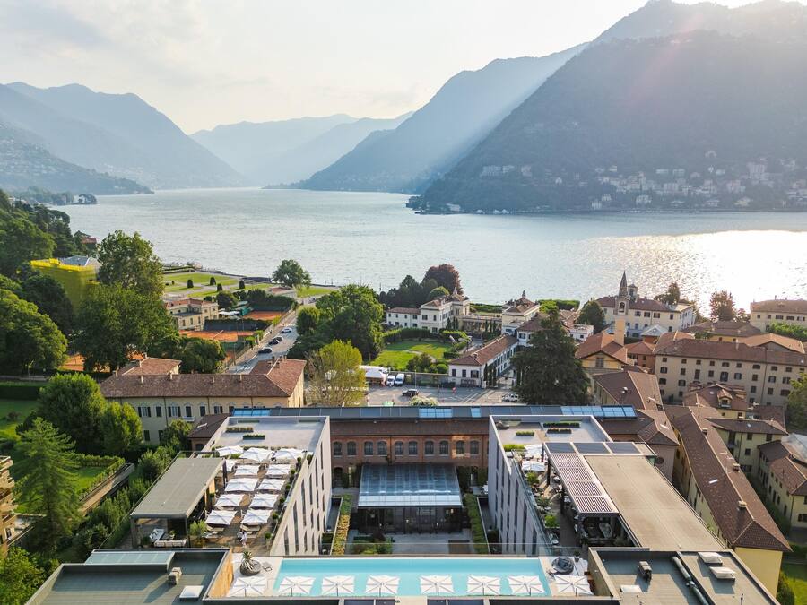A hotel with a rooftop pool, surrounded by lush greenery and a view of a lake and mountains.