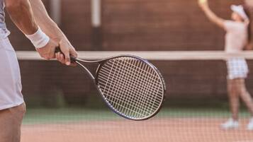 A tennis court with a net in the center, a player in the foreground holding a racket, and another player in the background preparing to serve.