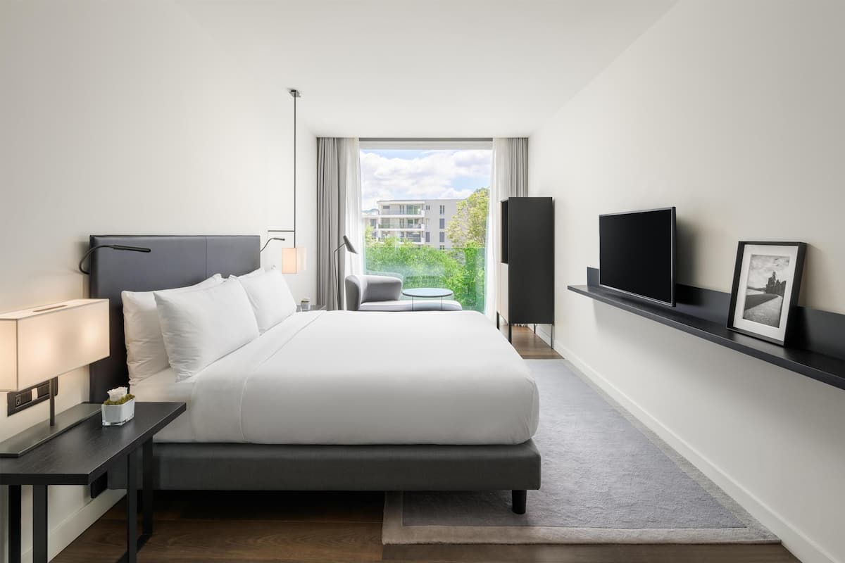 Une chambre d’hôtel moderne, dotée d’un grand lit, d’une table de chevet, d’une lampe, d’une télévision et offrant une vue sur la verdure environnante.