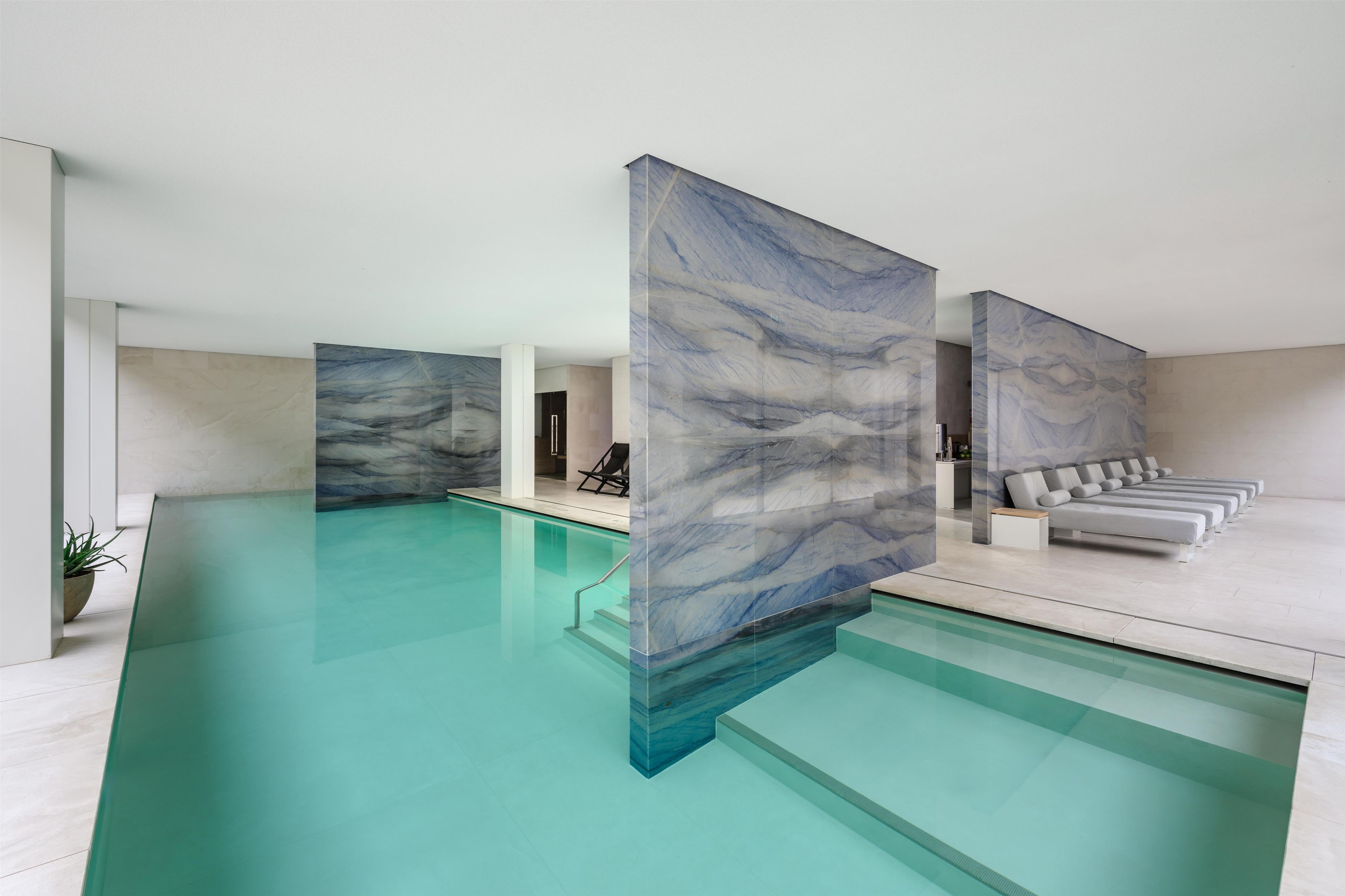 A modern indoor pool area with a clear pool, marble wall panels, and a seating area with a sofa and chairs.