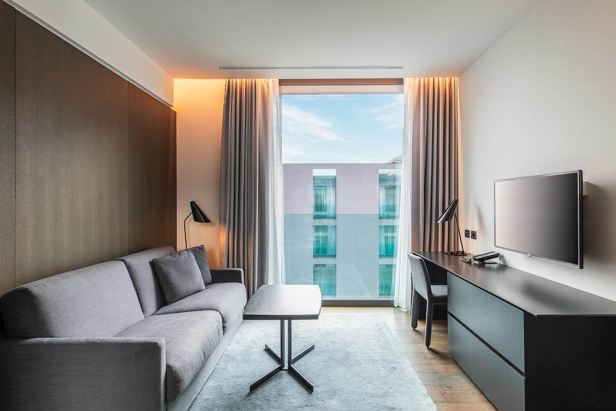 Une chambre d’hôtel moderne équipée d’un canapé, d’une petite table, d’un bureau, d’une télévision et d’une grande fenêtre avec des rideaux.