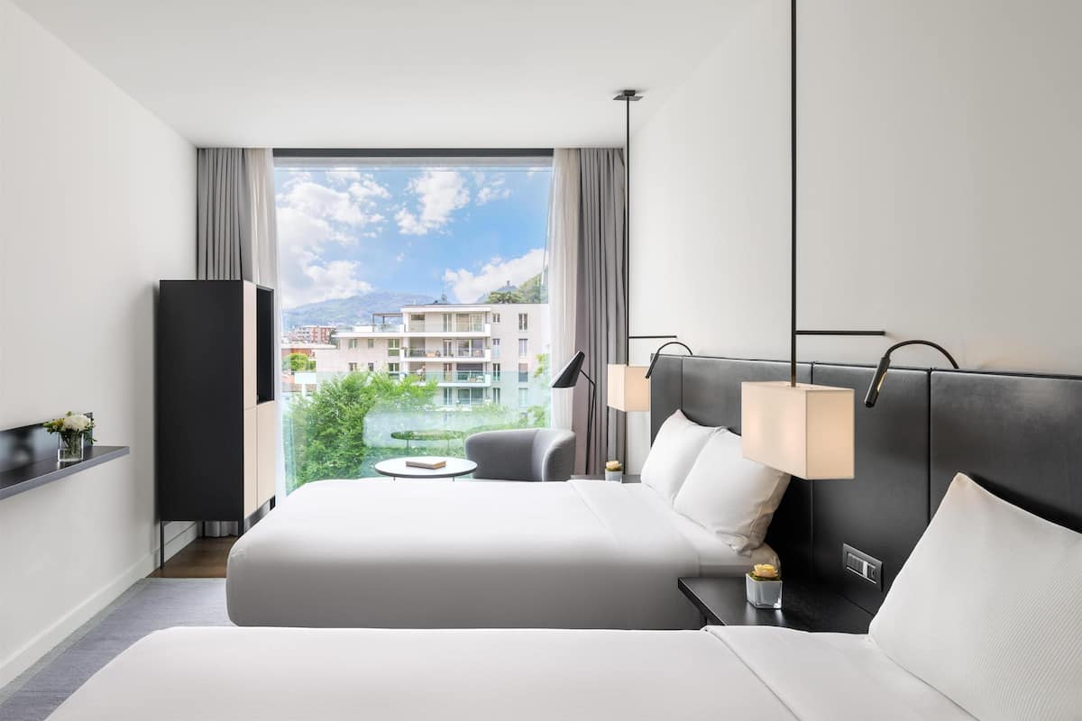 Une chambre d’hôtel avec un grand lit, des lampes de chevet, une petite table, un téléviseur et une vue sur des bâtiments et des espaces verts.