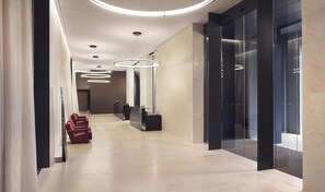 A modern hallway with a reception desk, red chairs, and a glass elevator.