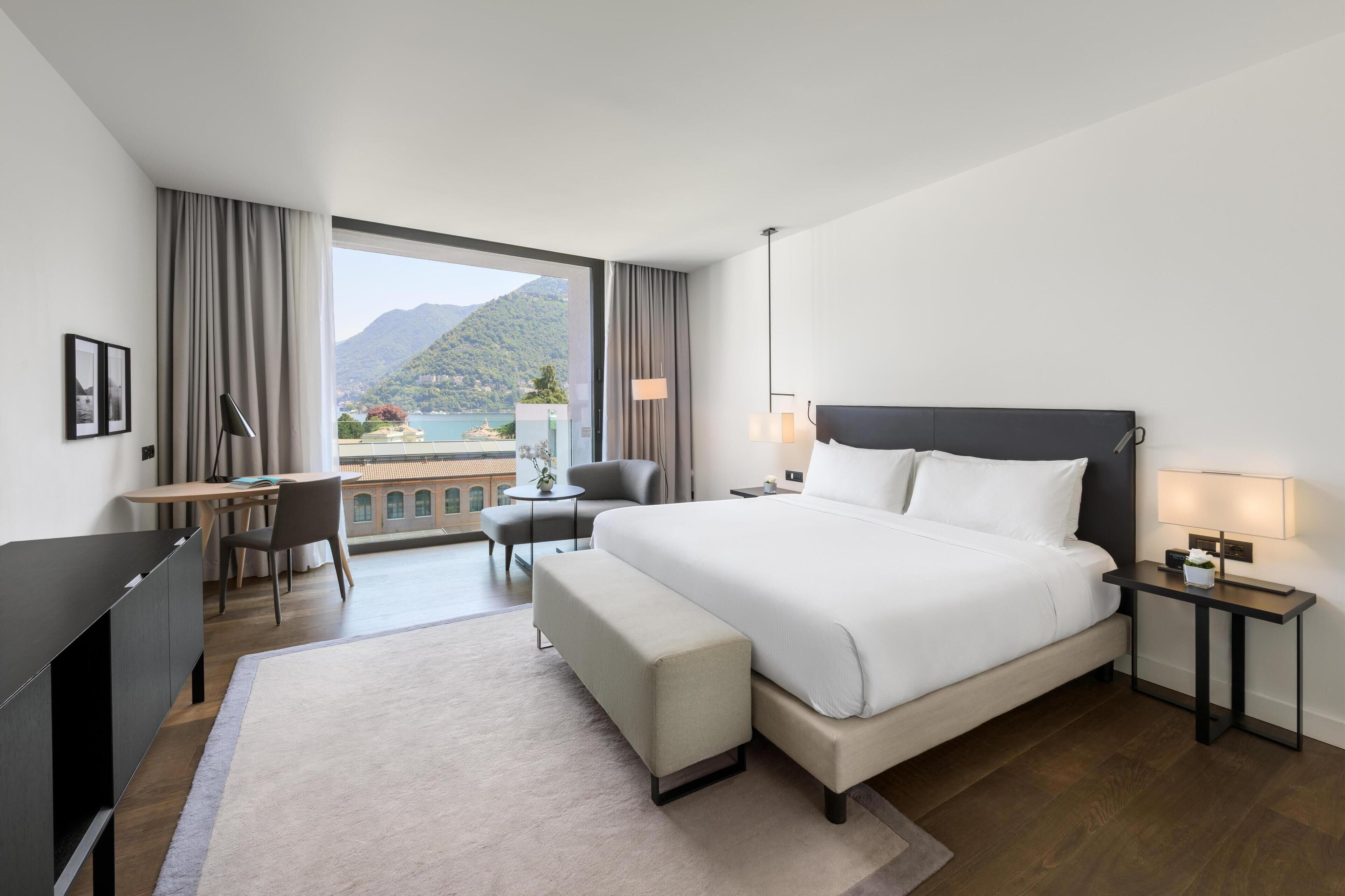 Une chambre d’hôtel avec un grand lit, un bureau, une chaise, une petite table et une vue sur les montagnes et un lac.