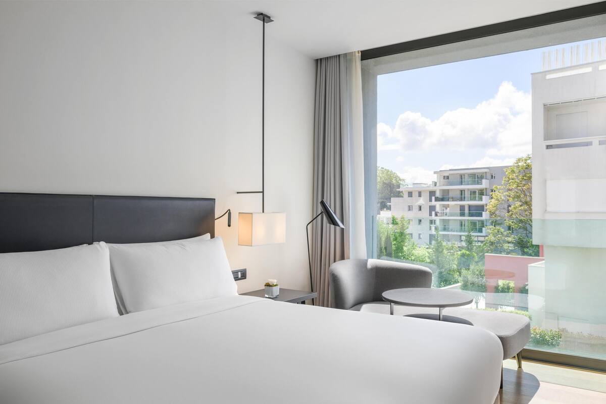 Une chambre d’hôtel avec un grand lit, une petite table, une chaise, une lampe et une vue sur les bâtiments par une grande fenêtre.