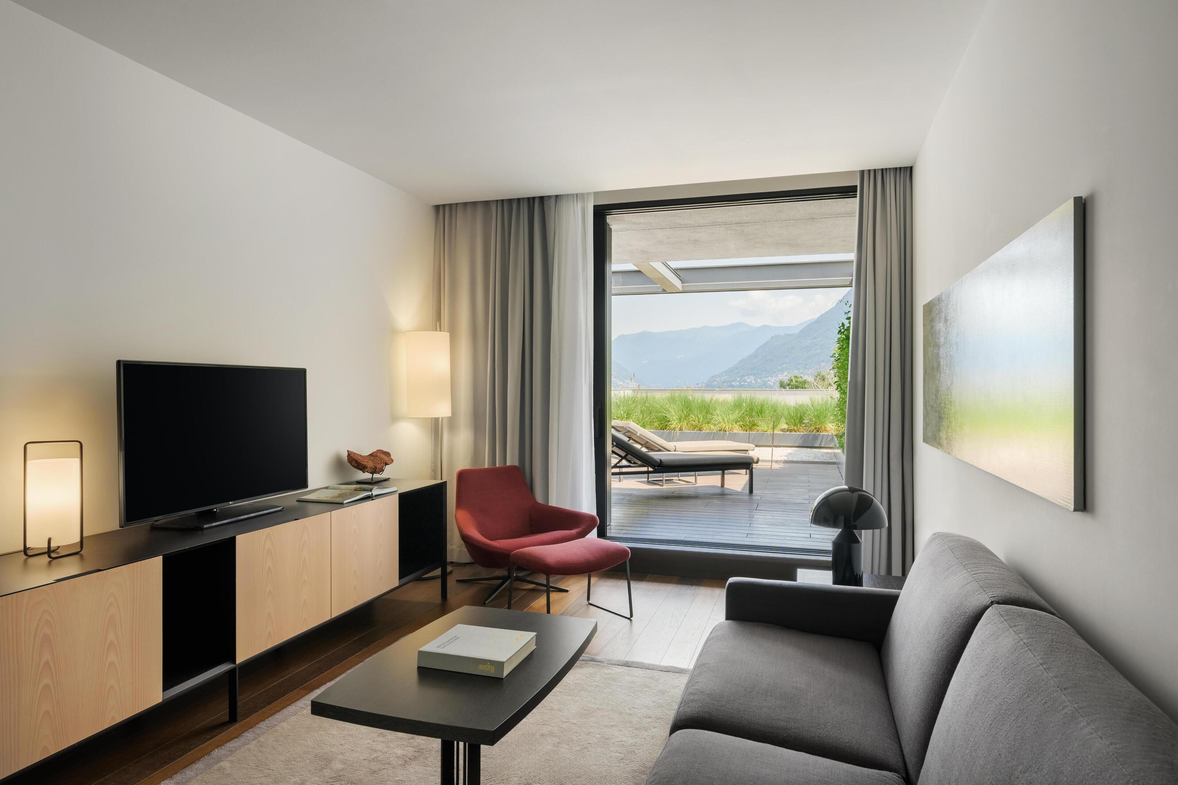 A modern hotel room with a flat-screen TV, a red armchair, a grey sofa, a coffee table, and a large window offering a view of mountains and greenery.