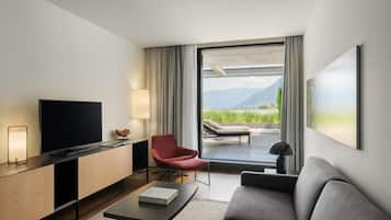 Ein modernes Hotelzimmer mit Flachbildfernseher, einem roten Sessel, einem grauen Sofa, einem Couchtisch und einem großen Fenster mit Blick auf Berge und Grün.