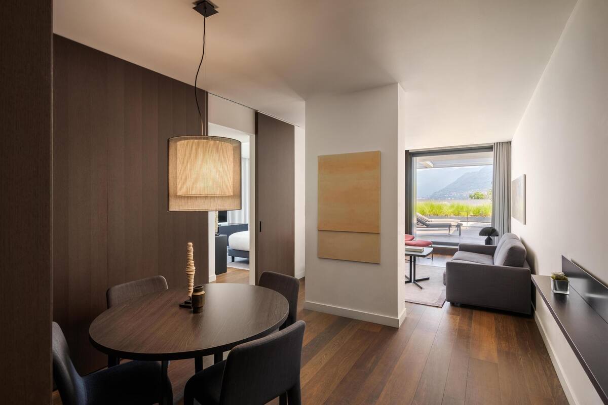 Un intérieur moderne avec une table à manger ronde, des chaises, une lampe suspendue, un canapé et une grande fenêtre donnant sur un paysage.