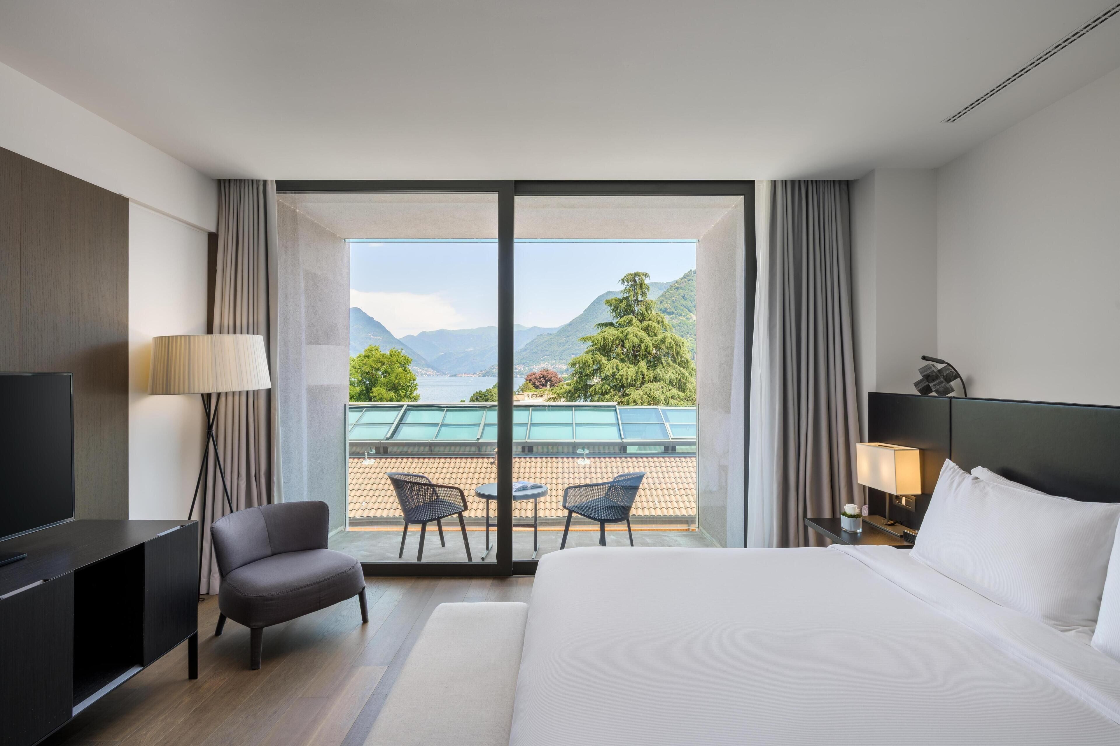 Habitación de hotel con cama, televisor, escritorio, silla, lámpara y vistas a un balcón con mobiliario exterior y un paisaje pintoresco.