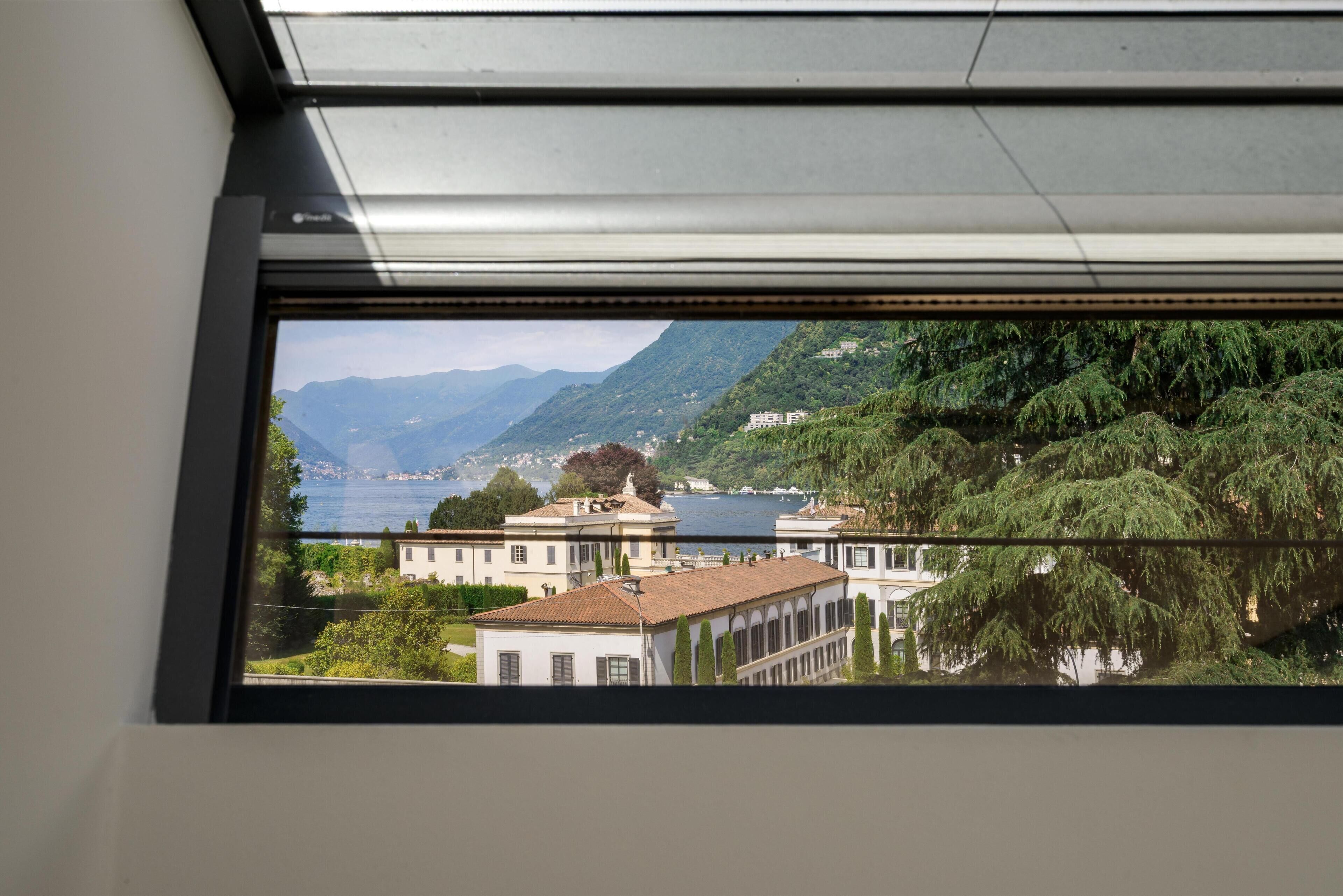 A view through a window of a building with a terrace, a body of water, and mountains in the background.