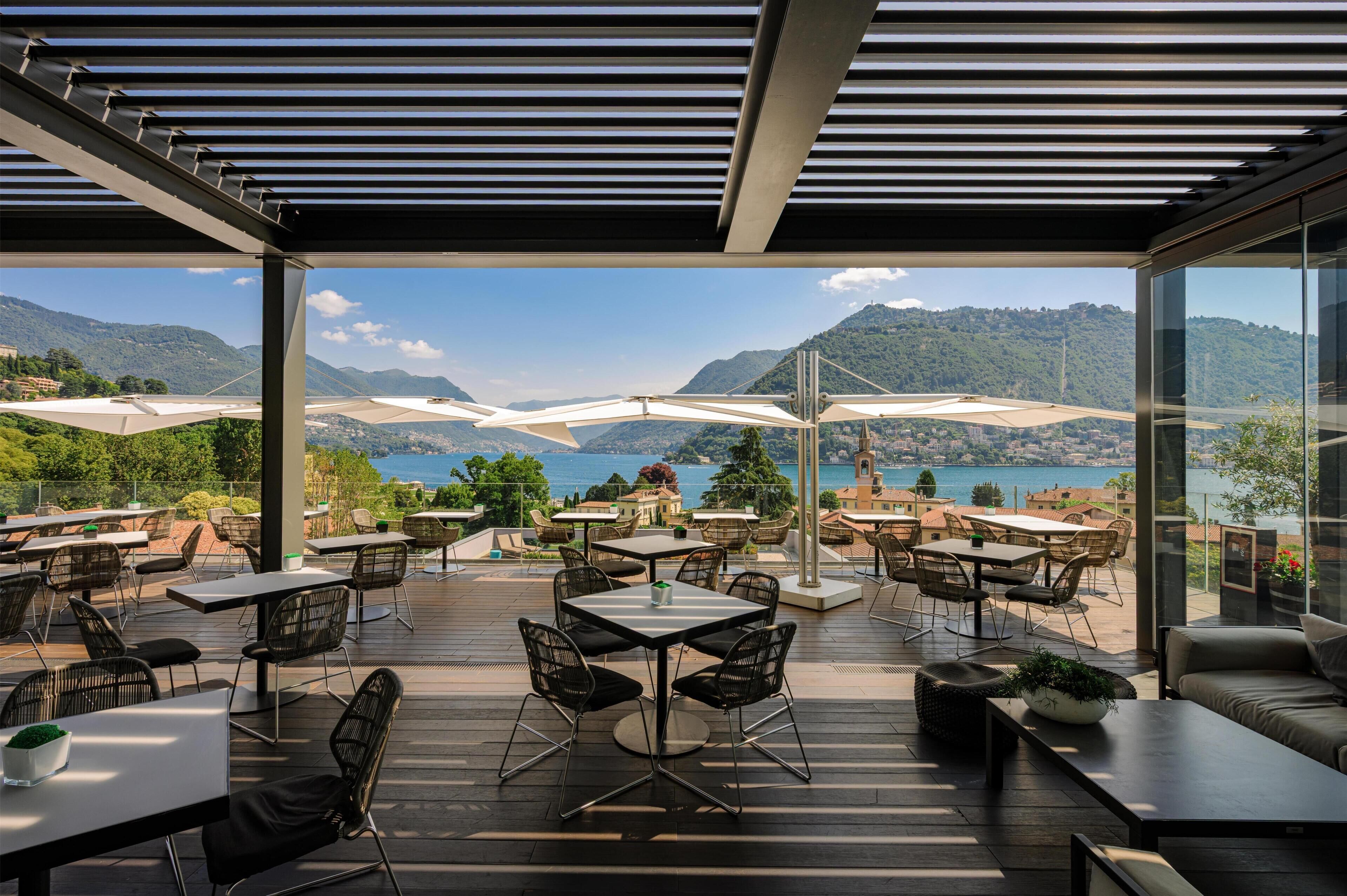 Un espace de restauration en plein air, avec des tables et des chaises, offrant une vue sur un paysage montagneux et une étendue d’eau.