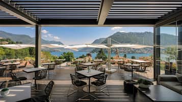 Un espace de restauration en plein air, avec des tables et des chaises, offrant une vue sur un paysage montagneux et une étendue d’eau.