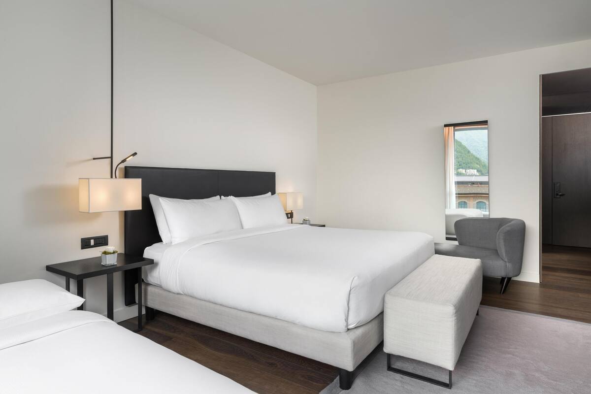 Une chambre d’hôtel moderne avec un grand lit, des tables de chevet, une chaise et une vue sur un immeuble par la fenêtre.