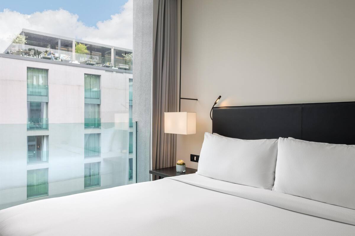 Une chambre d’hôtel avec un grand lit, une table de chevet avec une lampe, une vue sur un immeuble à travers une paroi en verre et un balcon avec des sièges d’extérieur.