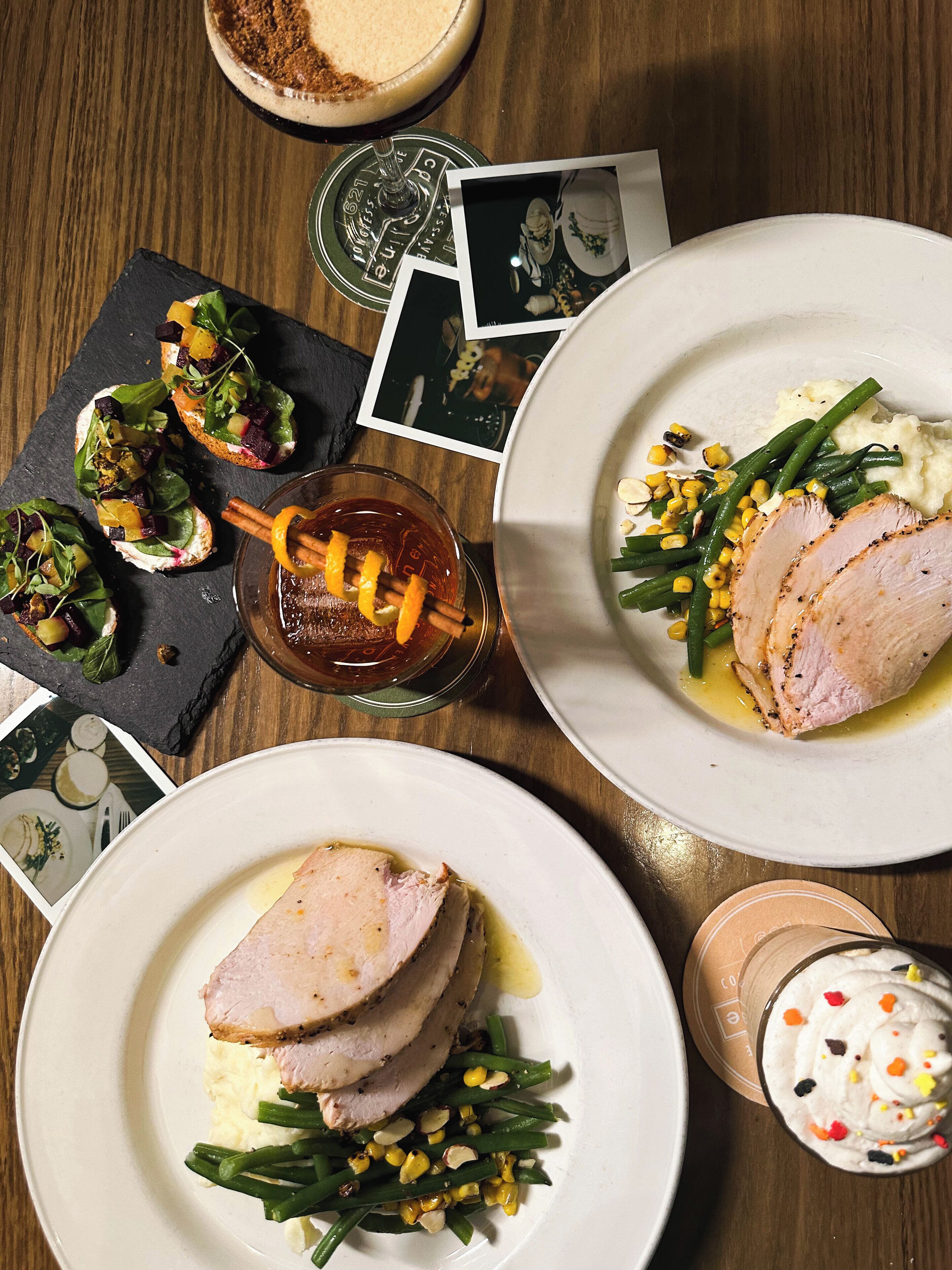 A dining table with plates of food, including sliced meat, green beans, and corn. There are also photos and a glass of drink on the table.