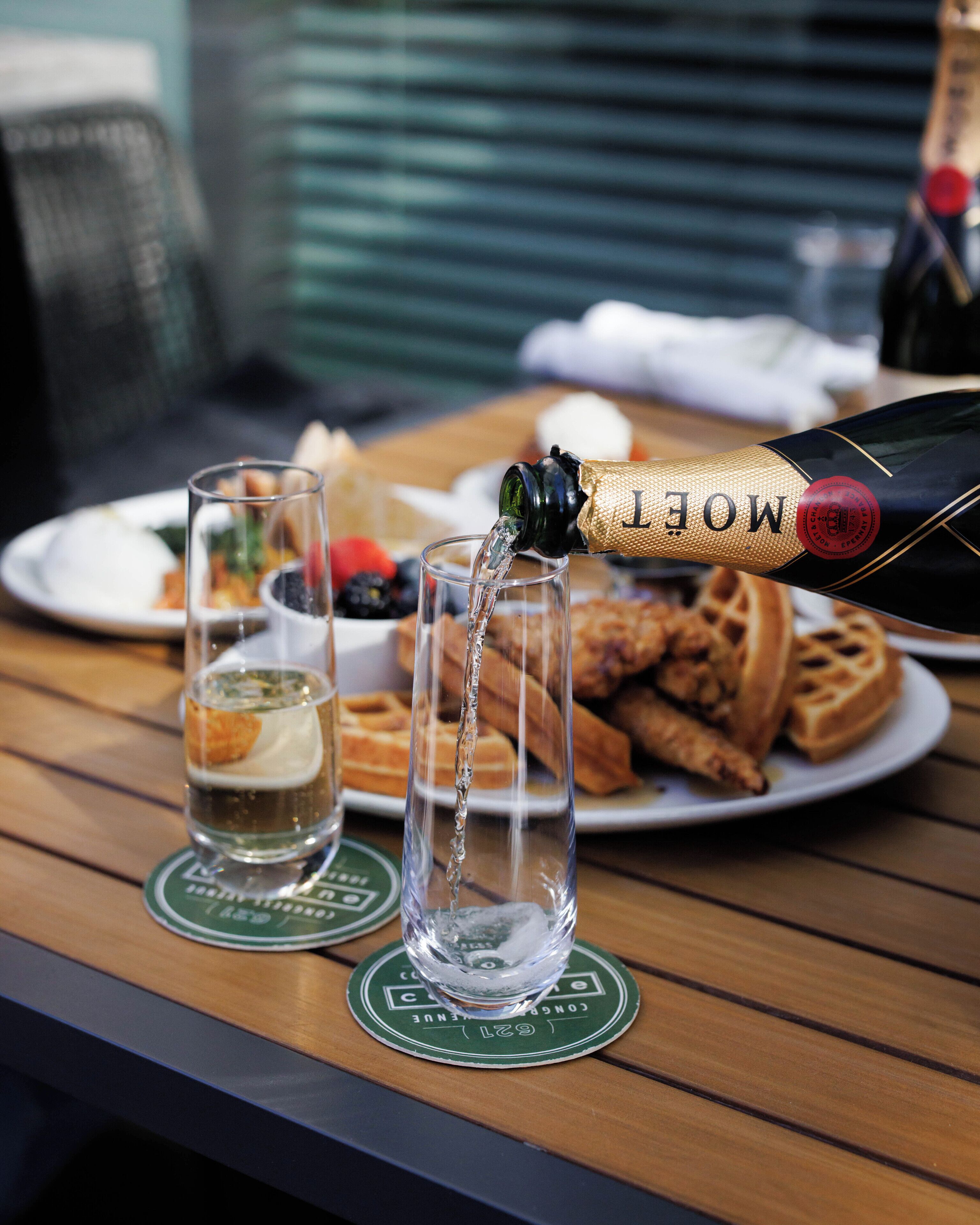 Una botella de Moët champagne siendo servida en una copa sobre una mesa de madera, con gofres y un plato de comida al fondo.